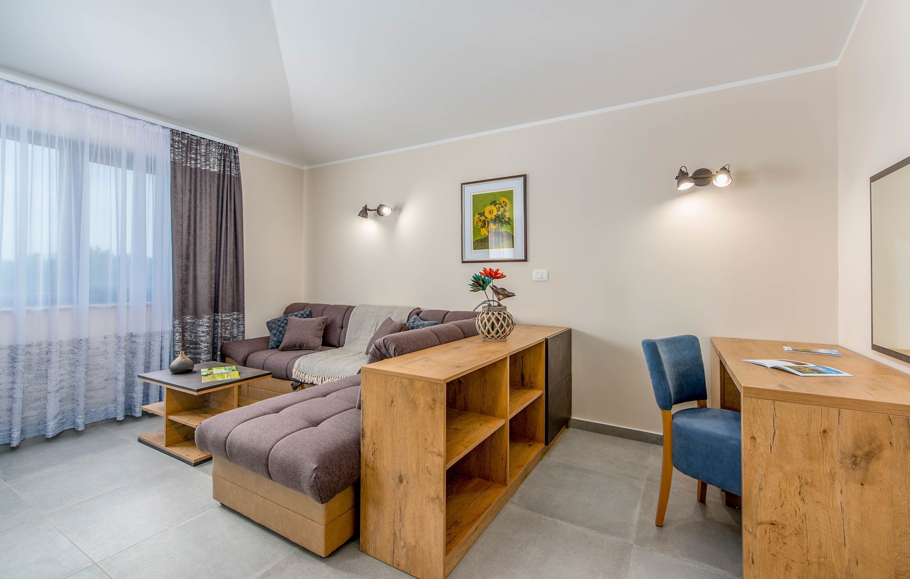 Una sala de estar moderna con un sofá, una mesa de centro y un escritorio. Hay una ventana con cortinas translúcidas, una lámpara de pared y un cuadro en la pared.