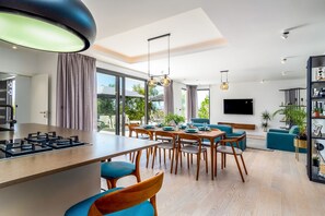 A modern kitchen with a central island, dining area with chairs, a living room with a sofa, and a TV mounted on the wall.