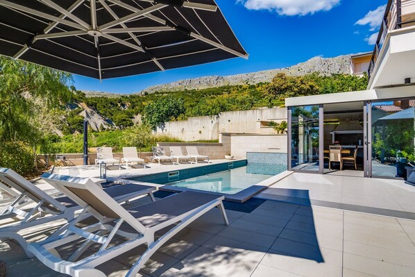 A modern outdoor pool area with lounge chairs, a glass-enclosed dining area, and a view of a mountainous landscape.