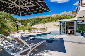 A modern outdoor pool area with lounge chairs, a glass-enclosed dining area, and a view of a mountainous landscape.