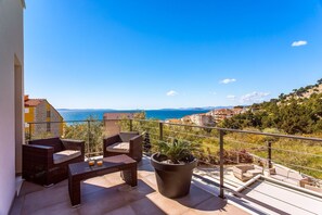 Ein Balkon mit Korbmöbeln, einer Topfpflanze und Blick auf das Meer und Gebäude.