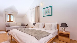 Ett sovrum med en stor säng, en mindre säng, ett nattduksbord, en lampa, en tavla och ett fönster med gardiner.