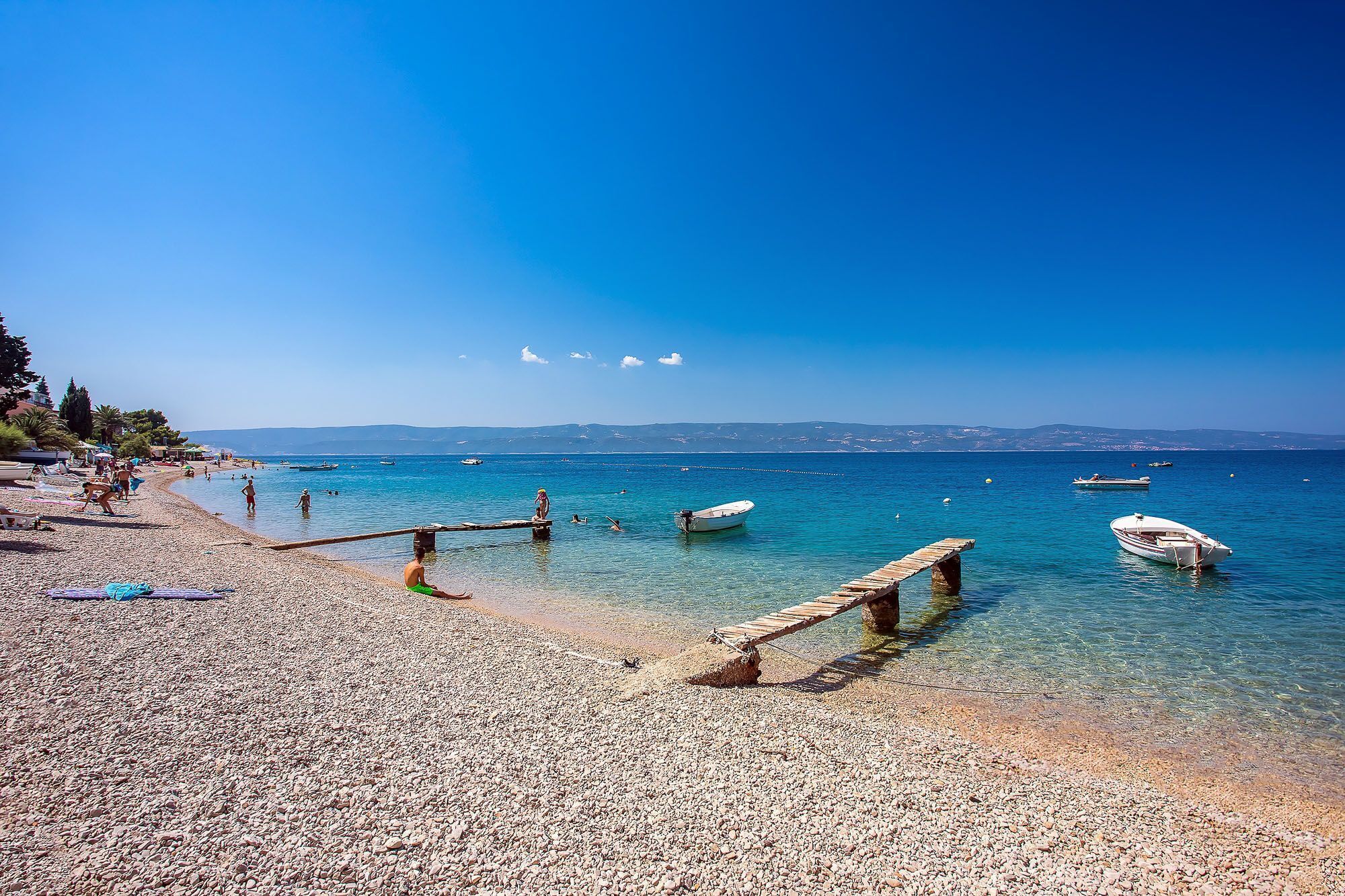 A pebbled beach with boats, clear water, and a wooden pier.