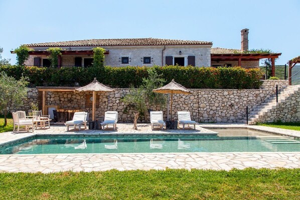 Un espace piscine avec des chaises longues et des parasols, un mur en pierre et, au fond, un bâtiment avec un toit en tuiles.