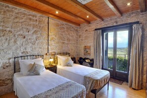 Une chambre avec deux lits, des murs en pierre, des poutres en bois au plafond et une grande fenêtre avec des rideaux.