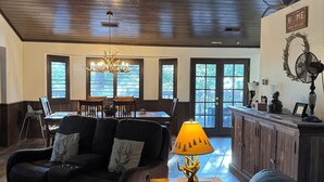 Una sala de estar acogedora con techo de madera oscura, un sofá de cuero negro, un comedor con mesa y sillas, y una lámpara decorativa.