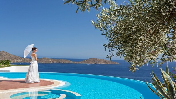 A person in a white dress standing by a pool with a view of the sea and mountains.