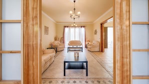 A spacious living room with a central coffee table, two sofas, a chandelier, and a large window with curtains. - Villa Kalvos - Five Bedroom Villa, Sleeps 10 (Kolympia)