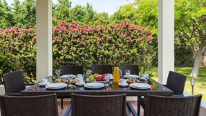 An outdoor dining area with wicker chairs and a table set for breakfast, surrounded by lush greenery and flowering bushes. - Villa Kalvos - Five Bedroom Villa, Sleeps 10 (Kolympia)