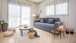 A living room with a grey sofa, a round coffee table, and a small wooden side table. There are two potted plants, a wicker basket, and a floor lamp.
