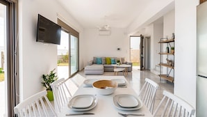 Un espace de repas moderne avec une longue table dressée pour quatre personnes, un canapé en arrière-plan et un téléviseur fixé au mur.