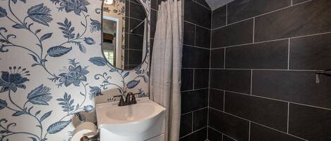 Un baño con lavamanos blanco, un espejo y una zona de ducha con azulejos oscuros.