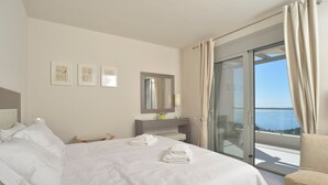 A hotel room with a bed, a desk, a mirror, and a view of the sea through a sliding glass door. - Villa Ipsos - Five Bedroom Villa, Sleeps 9 (Spartilas)