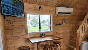 Interior de una cabaña de madera con una mesa, taburetes, una ventana y un equipo de aire acondicionado.
