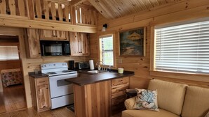 Una cocina acogedora de estilo cabaña, con paredes de madera, estufa, microondas y lavamanos. Hay una ventana con persianas y un cuadro en la pared.