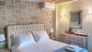 Ein Schlafzimmer mit Steinwand, einem gepolsterten Kopfteil, einem Bett mit weißen Leinen, einem Nachttisch mit Lampe, einer Kommode mit Spiegel und einer kleinen Pflanze.