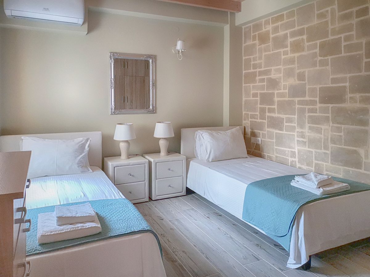 A hotel room with two beds, a small table, two bedside tables with lamps, a wall-mounted light, and a stone wall.