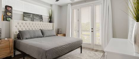 Un dormitorio con cabecera capitonada, cama con ropa de cama gris, ventilador de techo, mesita de noche blanca, una mesita auxiliar, un reloj de pared, un cuadro enmarcado y una ventana con persianas.