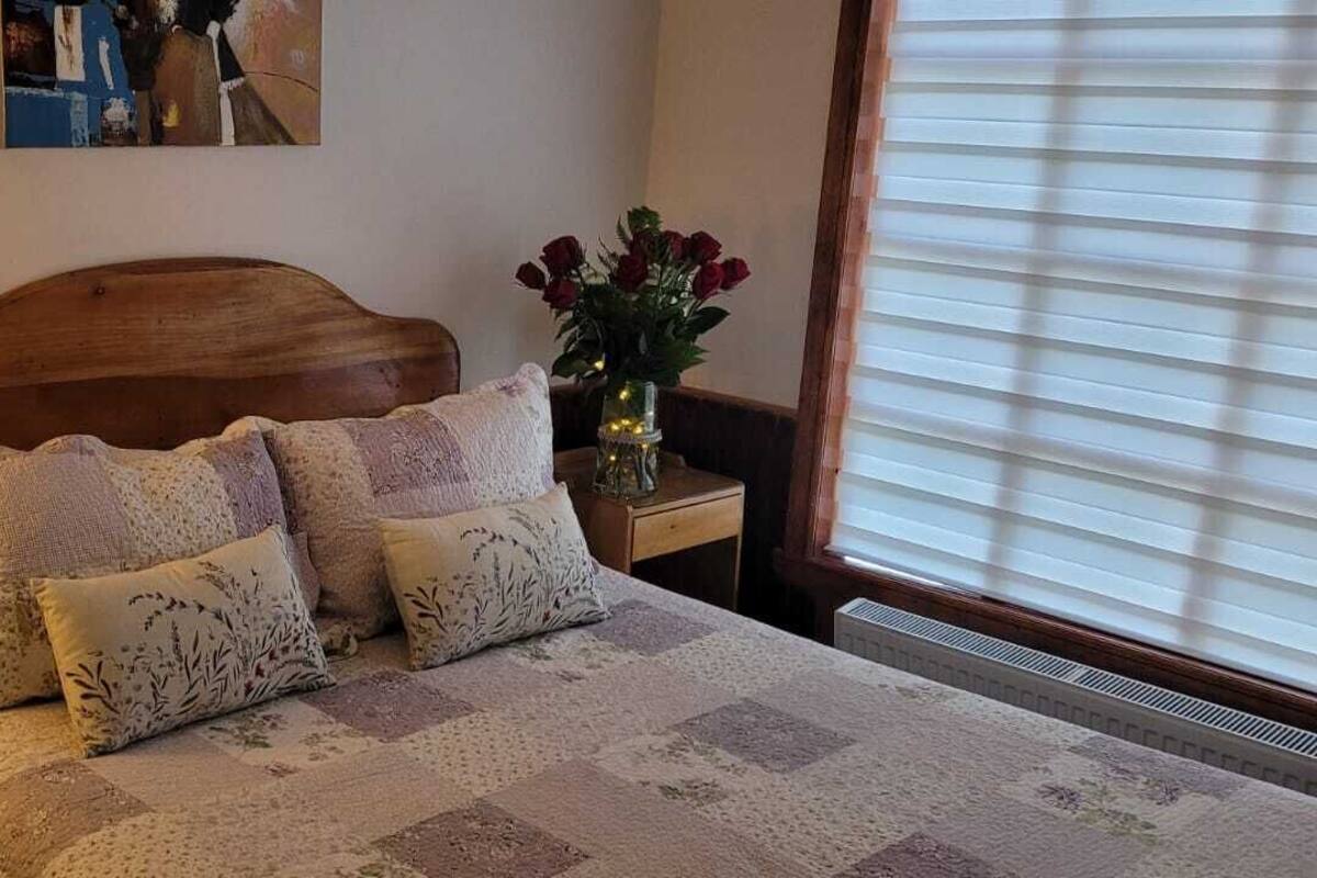 A neatly made bed with a quilt and two decorative pillows, a bedside lamp, a small table with a vase of flowers, and a painting on the wall.