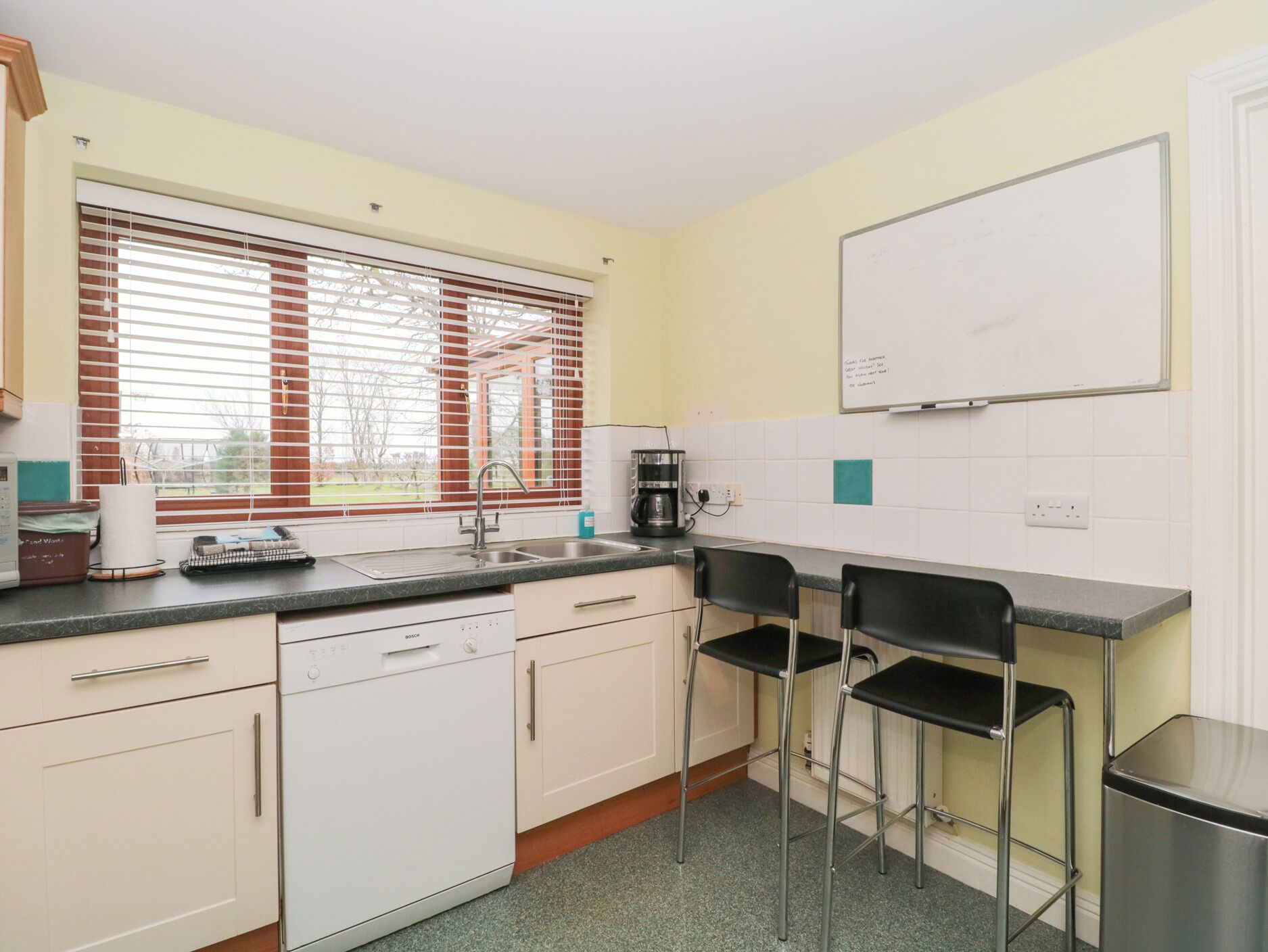 A kitchen with a sink, two bar stools, a coffee maker, and a whiteboard on the wall.