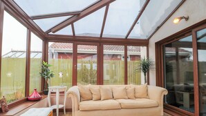 A conservatory with a wooden ceiling, a beige sofa, a small table with a lamp, and a yellow stool. - Morgan House (Bridgwater)