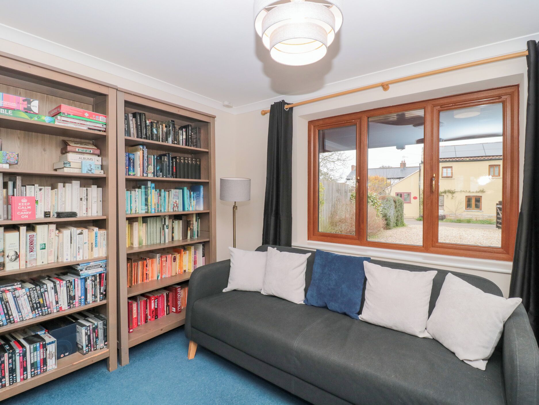 Ein gemütliches Zimmer mit grauem Sofa, zwei Bücherregalen voller Bücher, einem Fenster mit Vorhängen und einer Lampe.