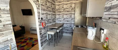 Una cocina compacta con protector mural de piedra, una zona de comedor con taburetes de bar y una pequeña sala de estar con sofá y televisor.