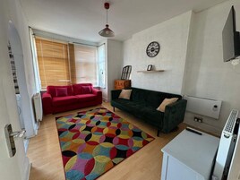A living room with a red and green sofa, a colorful rug, a wall clock, and a TV.