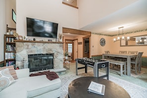 A living room with a stone fireplace, a flat-screen TV, a foosball table, a dining table with chairs, and a bookshelf.
