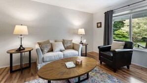 A living room with a sofa, a coffee table, a side table, a lamp, a chair, and a window with a view of greenery. - Spacious , RV parking, yard, cozy fireplaces, game library, near Manchester (Port Orchard)