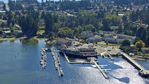 An aerial view of a marina with boats docked, a building with a large parking lot, and surrounding residential areas. - The Spot at Porpoise Bay (Sechelt)