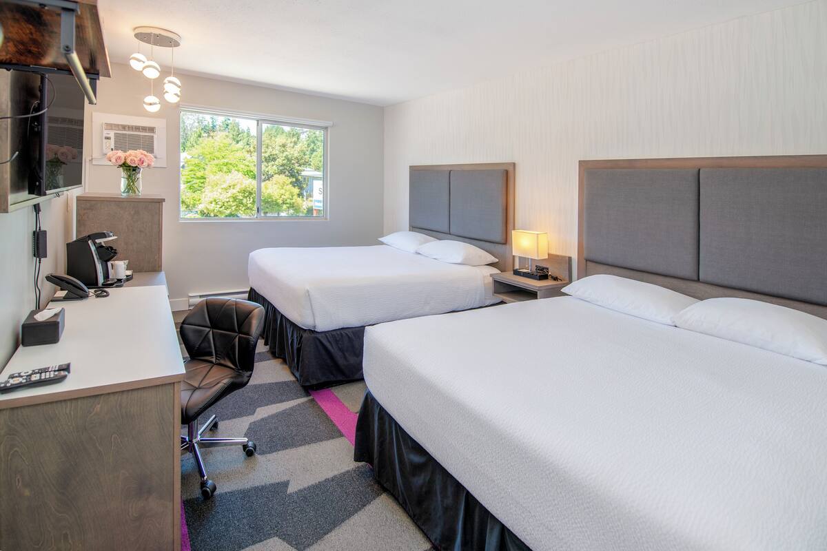 Habitación de hotel con dos camas, un escritorio con computadora, una silla y una ventana con vista a árboles.