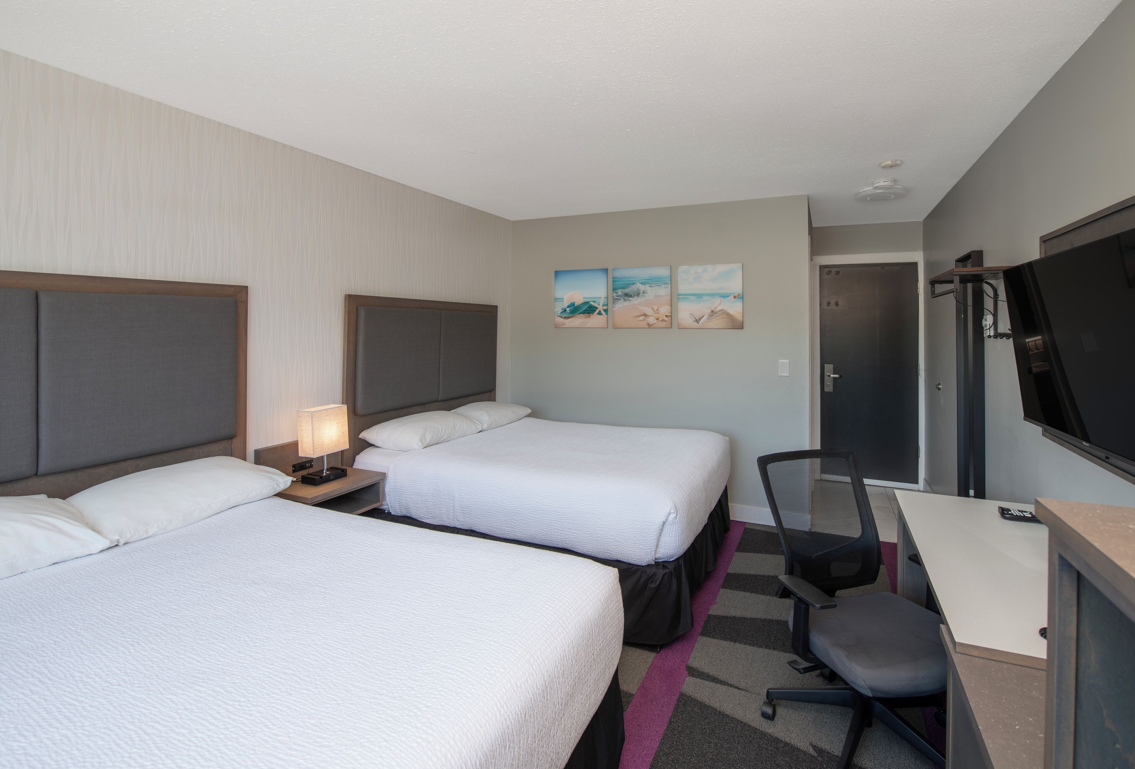 A hotel room with two beds, a desk, a chair, a TV, and artwork on the wall.