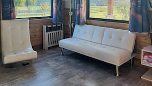 Une pièce avec un canapé blanc, un fauteuil et un radiateur. Il y a deux fenêtres avec des rideaux, et une vue sur l’extérieur.