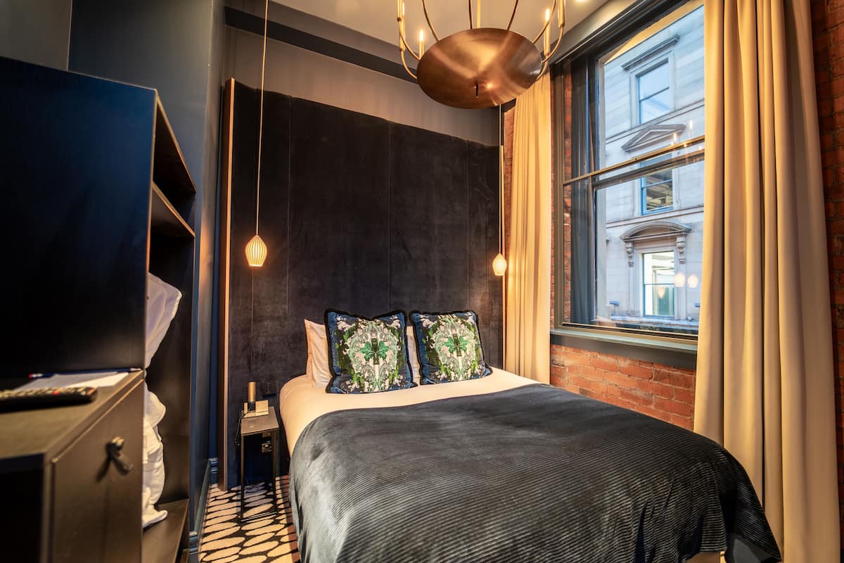 A modern hotel room with a large bed, a TV, and a view of the city through a large window.