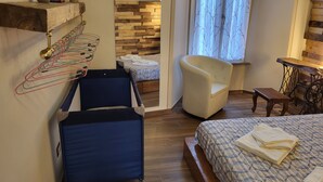 Un dormitorio con cama, una silla, una mesita pequeña y un estante de madera con un espejo y algunos objetos.