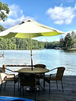 Una terraza de madera con una mesa y sillas bajo un paraguas grande, con vistas a un lago con árboles y un bote al fondo.