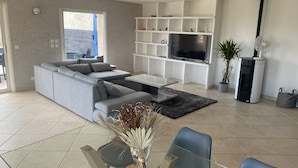 A modern living room with a grey sectional sofa, a glass coffee table, a TV on a white shelf, a fireplace, and a dining area with a table and chairs. - Spacious villa with pool in Nice (Nice)