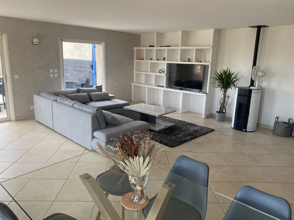 A modern living room with a grey sectional sofa, a glass coffee table, a TV on a white shelf, a fireplace, and a dining area with a table and chairs.