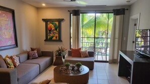 A living room with a sofa set, a coffee table, and a TV. There is a window with curtains and a view of palm trees. - Pacifico Luxury 3B/2B condo, 10 minutes walk to the beach (Playa Del Coco)