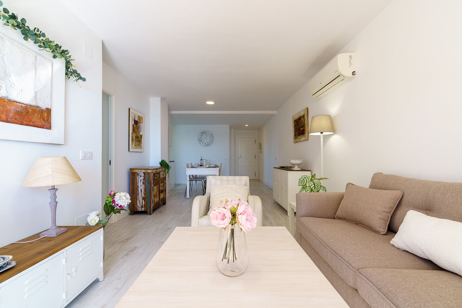 Una sala de estar moderna con un sofá, una mesa de centro y un florero. Se observa un comedor al fondo con una mesa y sillas.