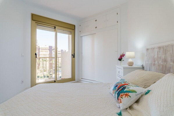 Un dormitorio con una cama grande, una mesita de noche con lámpara, un ventanal con vistas a edificios y una puerta corredera.