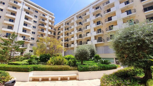 A multi-story residential building with balconies, surrounded by well-maintained greenery and a paved courtyard. - Centrally located and modern apartment CKNI1 (Gzira)