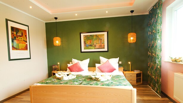 A bedroom with a bed, two bedside tables, a nightstand, a green wall, and two hanging lamps.