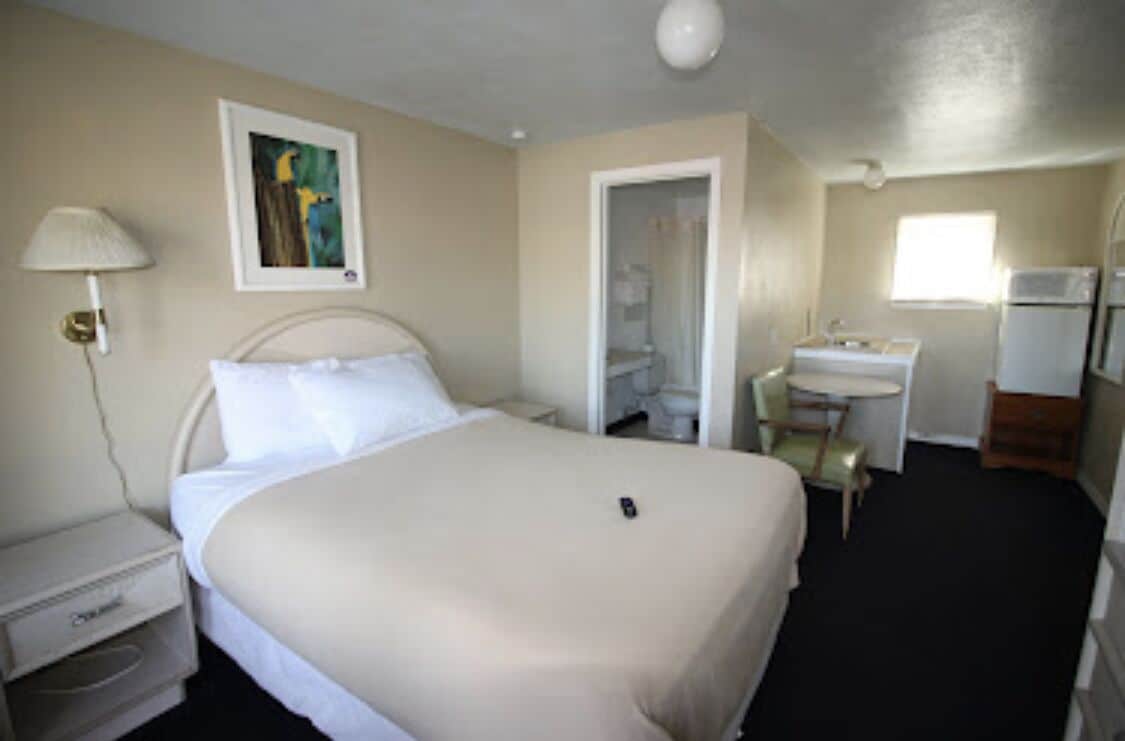 A hotel room with a bed, a desk, a chair, a lamp, and a refrigerator.