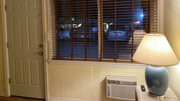 A room with a closed door, a window with blinds, a lamp, and an air conditioning unit. - Sunland Inn Motel (Ephrata)