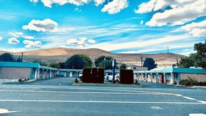 A motel with a parking lot, a row of rooms with a blue roof, and a mountain in the background. - Sunland Inn Motel (Ephrata)