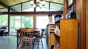 A wooden interior with a dining table and chairs, a bookshelf, and a large window.