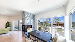 Una sala moderna con una mesa de comedor de madera oscura, asientos cómodos, chimenea y ventanas amplias con vista a la calle y al verdor.