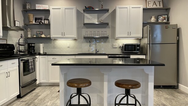 A modern kitchen with white cabinets, a stainless steel refrigerator, a microwave, and a double sink. There are two stools at the island. - Whispering Pines Guest House (Naples)